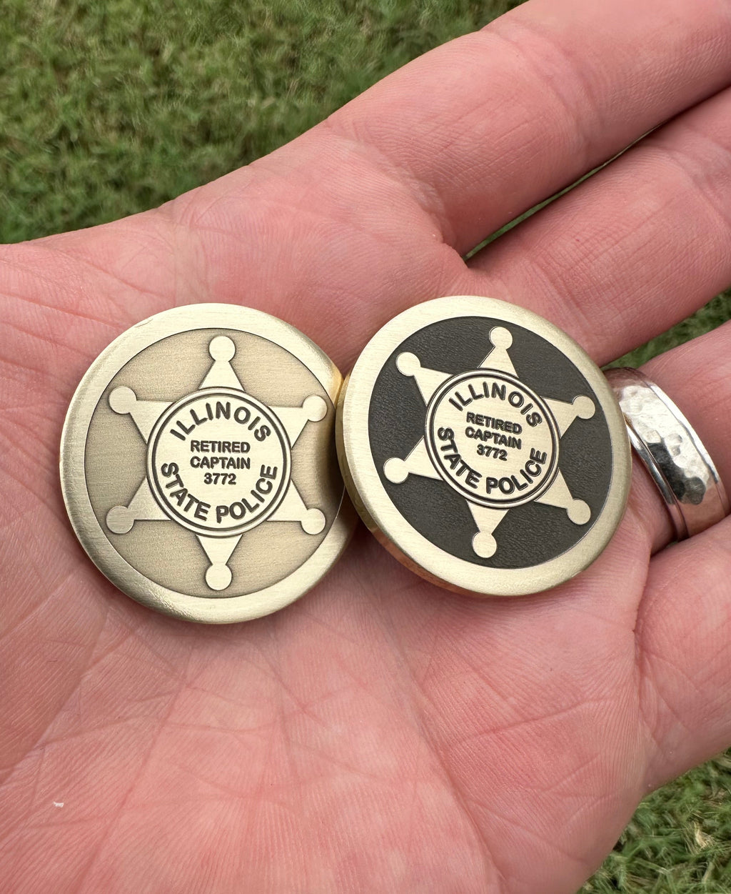 Two round brass ball markers with engraved designs held in a hand against a grassy background.
