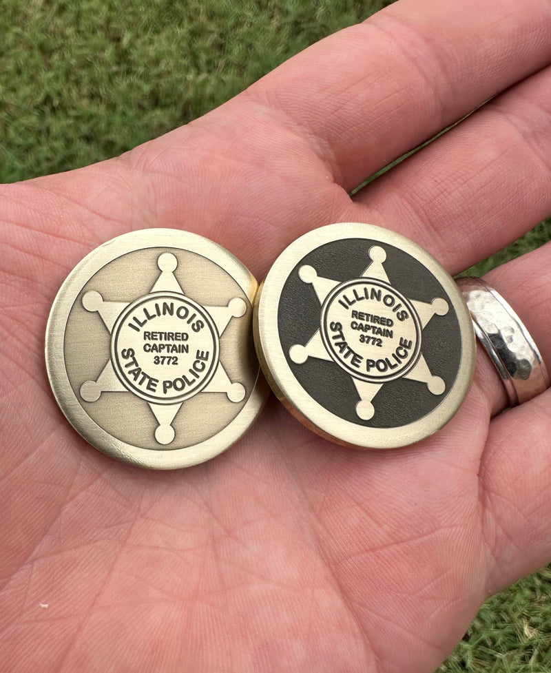Two round brass ball markers with engraved designs held in a hand against a grassy background.